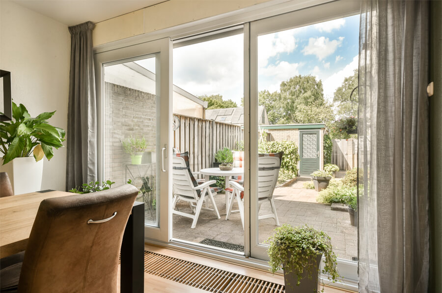 Un salon moderne comprend une chaise bleue, un divan beige et une télévision. De grandes portes-fenêtres mènent à une terrasse en bois avec des sièges extérieurs entourés d'arbres, mélangeant harmonieusement les espaces intérieurs et extérieurs.