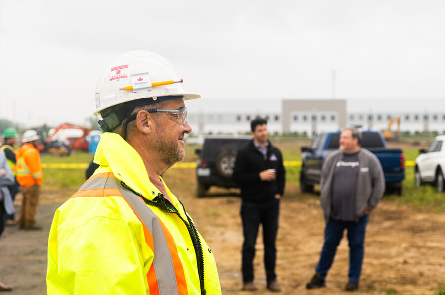 Un ouvrier de construction portant une veste de sécurité jaune et un casque blanc se tient au premier plan, souriant. À l’arrière-plan, des personnes discutent près de véhicules à une usine de fabrication de fenêtres à Ottawa (Ontario).