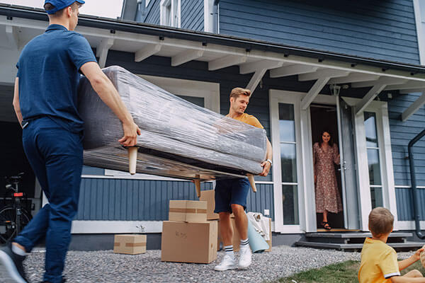 A couple men move a sofa into a home while a woman looks on from the front door.