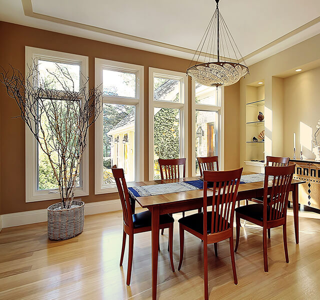 A dining room with a wooden table and six matching chairs. Large windows let in natural light, and a modern decorative chandelier hangs above. A potted tree stands in the corner next to built-in shelves displaying small items.