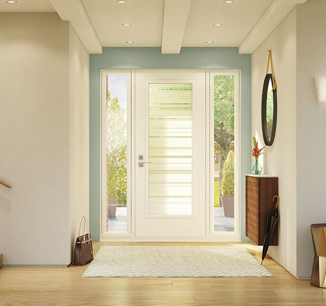 A modern, well-lit entryway features a white door with glass sidelites, a wooden side table with a red flower vase, and a round mirror on the wall. A striped rug lies on the light wood floor, with a brown bag placed next to the stairway.