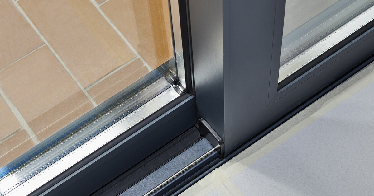 Close-up of a modern glass patio door with a metal frame. Part of a sliding mechanism, the floor features beige tiles inside and unfinished flooring outside. The well-lit scene highlights the sleek design and smooth sliding track.